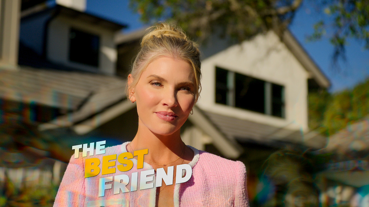 A woman with blonde hair in a pink jacket stands outside in sunlight with a suburban house in the background. Text on the image reads "The Best Friend." The setting is bright, with greenery and a blue sky.