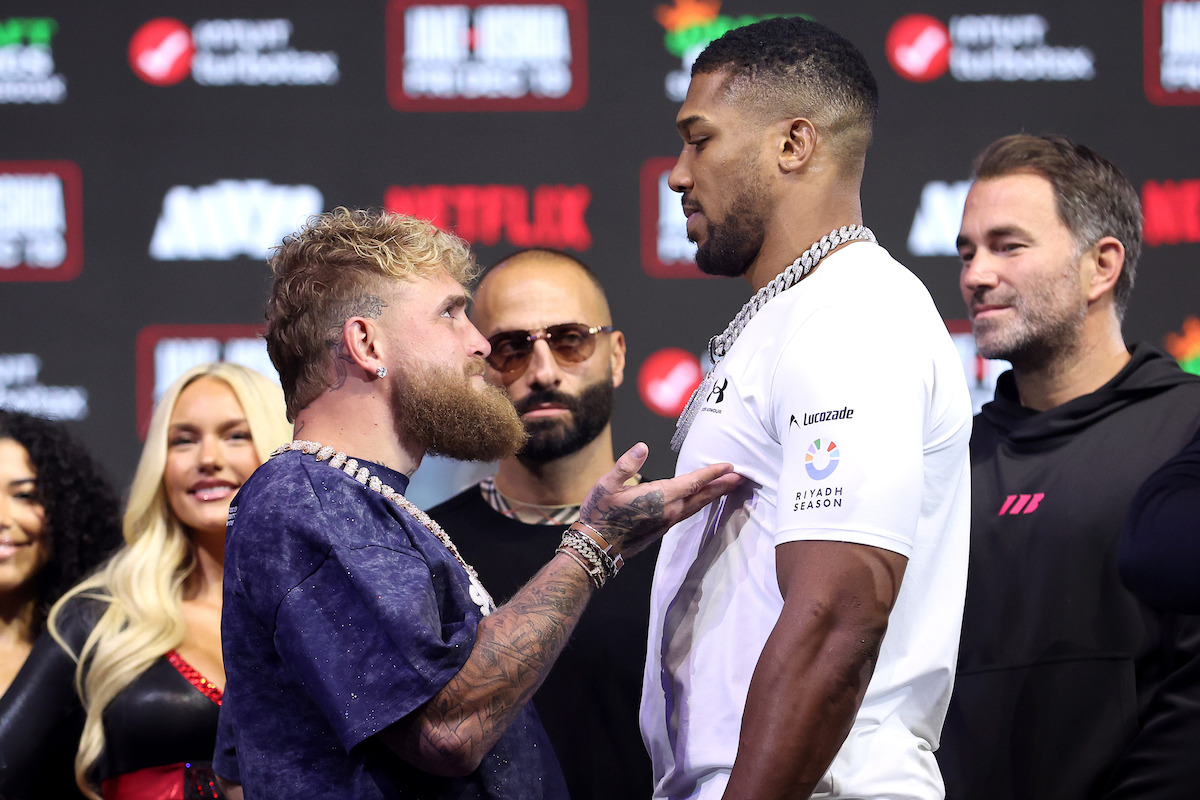 Jake Paul and Anthony Joshua face off on stage at a press event, surrounded by others and media backdrops, suggesting a pre-fight or sports promotion atmosphere with intense expressions and dramatic lighting.