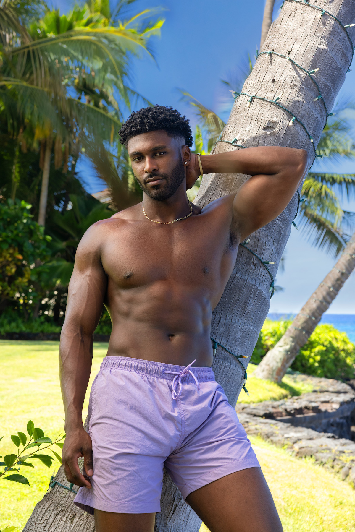 A man in lavender swim trunks stands by a palm tree at a tropical beachside location, with lush greenery, palm trees, and the ocean visible in the background under a clear blue sky.