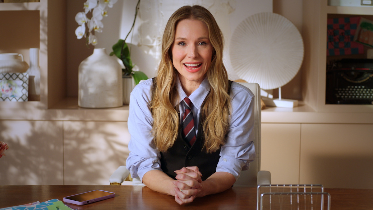 Kristen Bell sits at a desk in a well-lit office, wearing a vest and tie. Behind her are decorative vases, plants, papers, and a typewriter, creating a cozy, organized workspace.