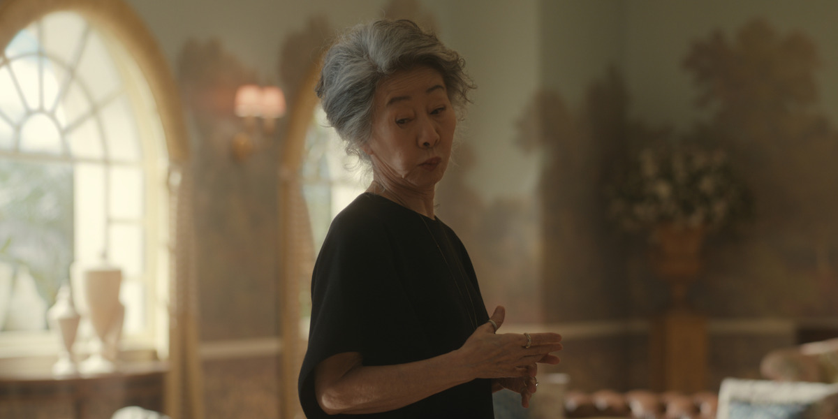 Older woman with gray hair in a black top stands indoors, softly lit elegant room with large arched windows, decorative wallpaper, classic furnishings, and floral arrangement in the background.