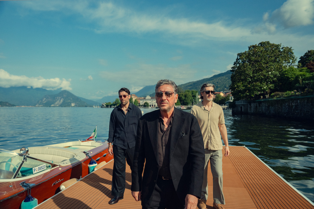 Three men in sunglasses walk on a wooden dock by a lake, luxury boats moored beside them, with scenic mountains and a village in the background under a blue sky with scattered clouds.