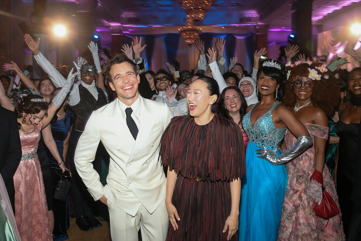 Two people pose and smile at the front of a lively group at a formal event, with guests in elegant dresses and suits, colorful lighting, and festive decorations in a ballroom setting.