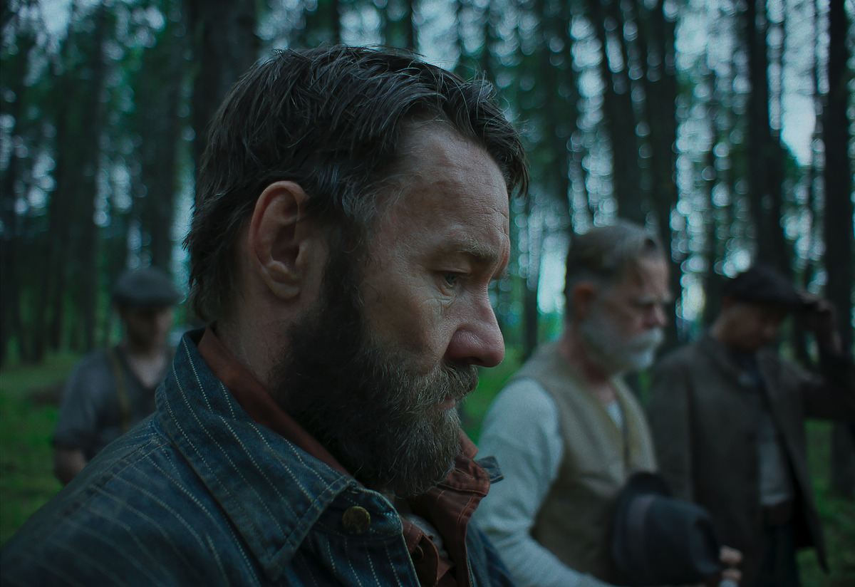 A bearded man in period clothing stands solemnly in a forest, surrounded by other serious-looking men in old-fashioned attire, creating a somber and reflective mood amidst the trees.