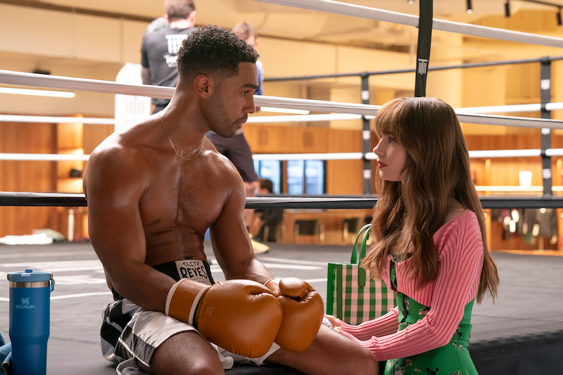  Lucien Laviscount as Alfie sits on the edge of a boxing ring while Lily Collins as Emily stands beside him in Season 4 of 'Emily in Paris'