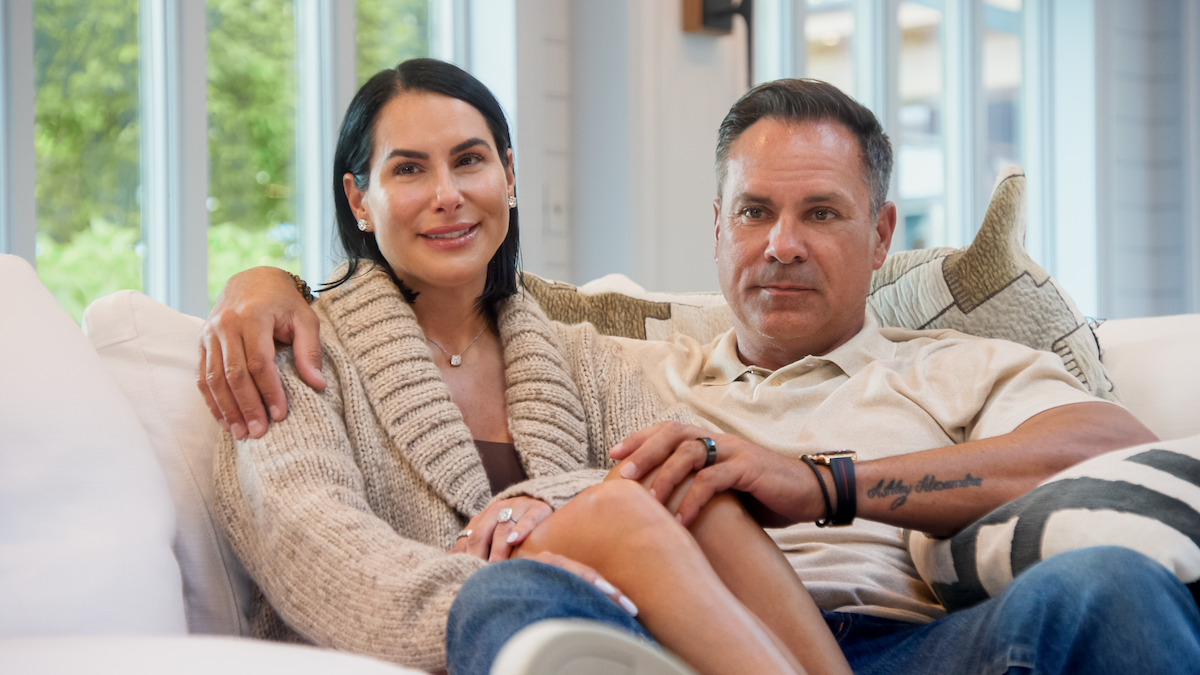 A couple sitting closely together on a white sofa in a bright living room, with large windows and green outdoor scenery in the background, looking relaxed and comfortable.