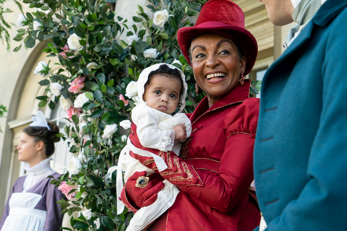 A woman in a red period costume holds a baby dressed in white, smiling outdoors near a rose-covered arch, with others in historical clothing in the background.