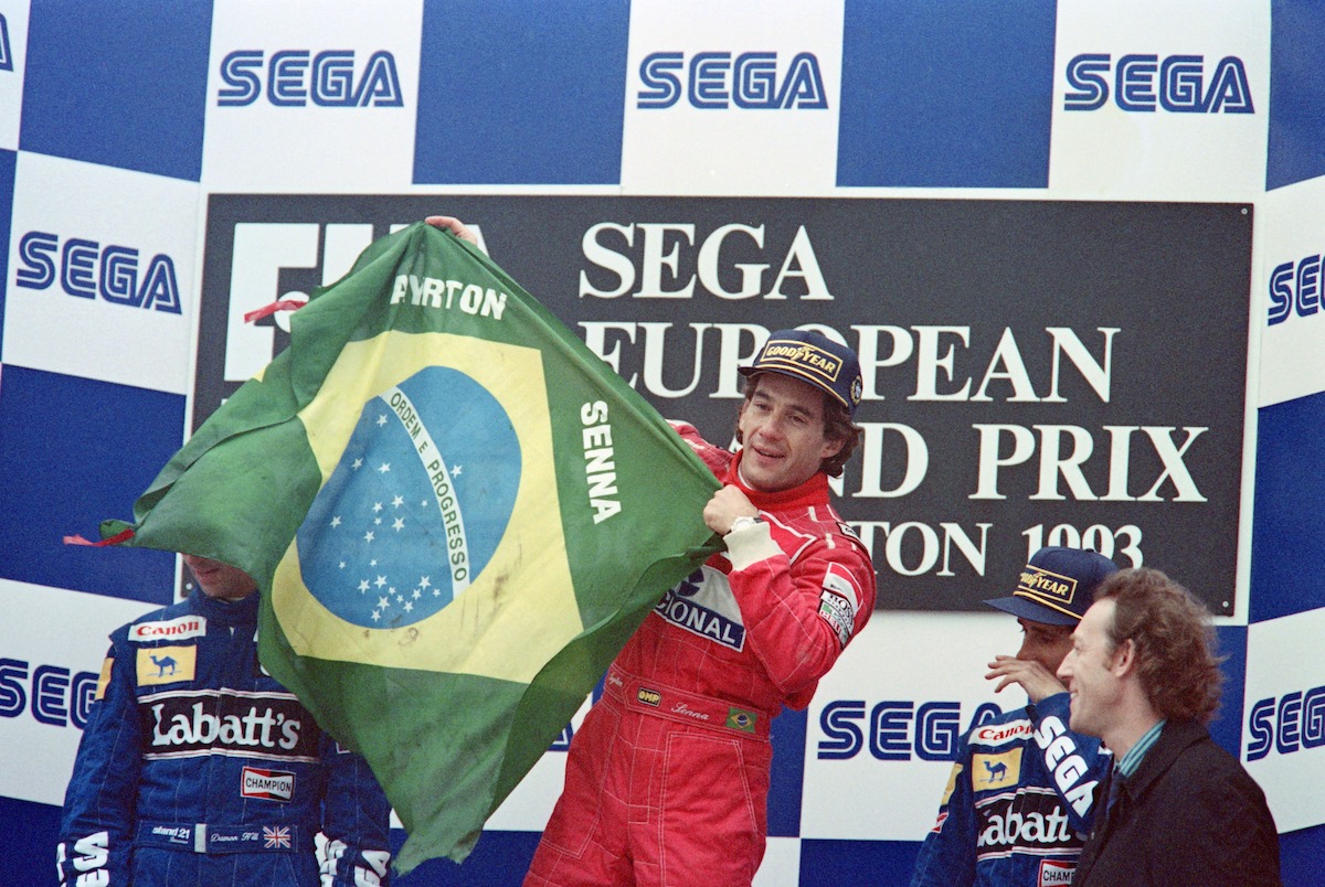 Ayrton Senna in his red racing suits stands on the top of the podium against a blue background that says "SEGA" as he waves a Brazilian flag with his name on it.