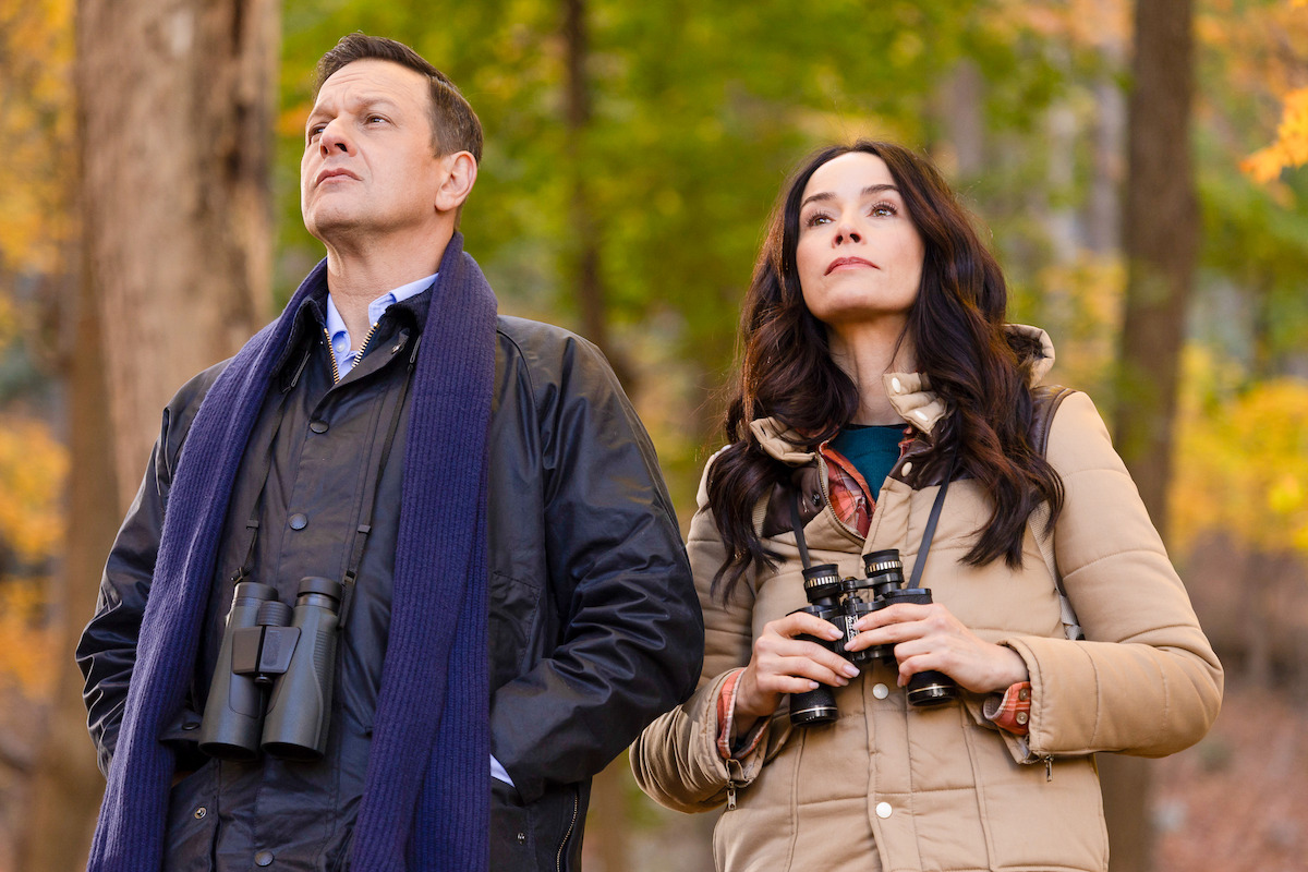 Two people in jackets hold binoculars while standing in a forest during autumn, looking up at something in the trees.