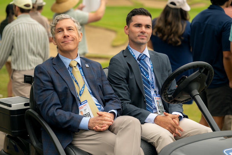 Two men in suits sit in a golf cart at a golf course event, wearing event badges, with several people standing in the background on a grassy area near sand traps. The setting appears formal and outdoors during daytime.
