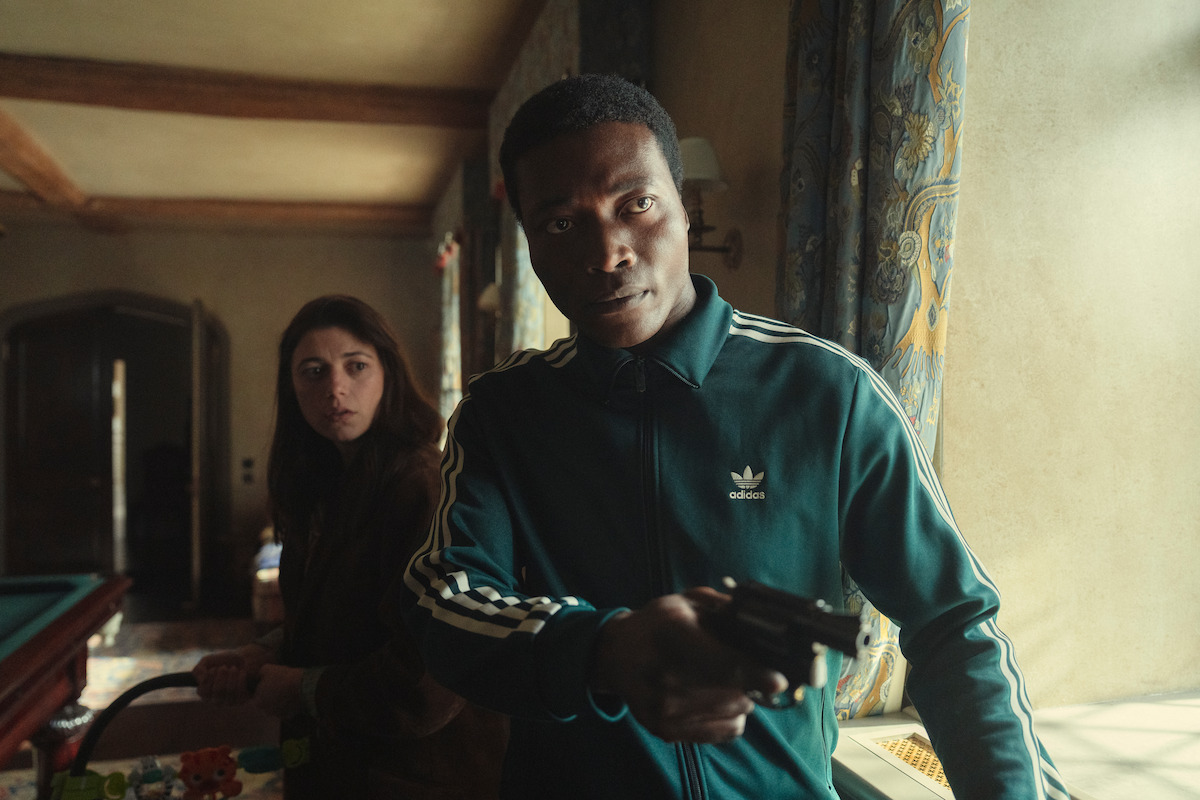 A man in a green tracksuit holds a gun in a dimly lit room, appearing tense, while a woman stands behind him looking concerned; patterned curtains and home interior details are visible.