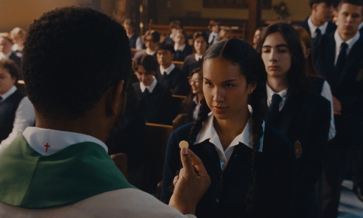 A priest offers communion to a girl in a school uniform during a church service, with other students in uniforms seated and standing in the background inside a church setting.
