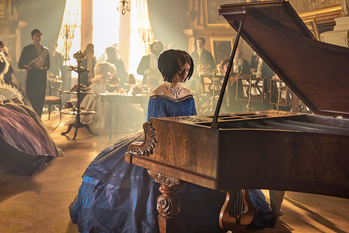 A woman in a blue Victorian dress plays a grand piano in an ornate, sunlit room with guests in period attire, creating an elegant and nostalgic atmosphere at a formal gathering.