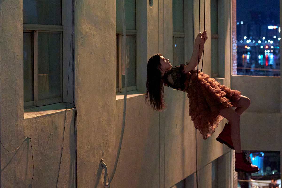 A woman in a ruffled dress and red boots hangs from ropes outside a tall building at night, with city lights reflecting on water in the background.
