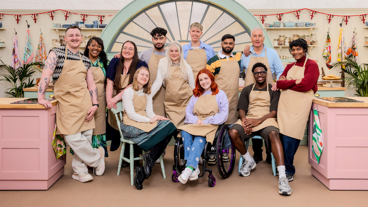 A diverse group of twelve people in aprons pose inside a colorful baking tent with pink counters, plants, and festive bunting, smiling for a group photo.