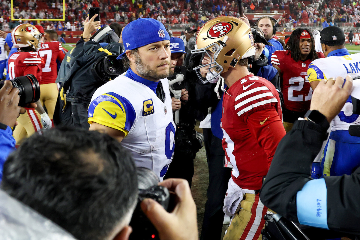 Two American football players from the Rams and 49ers face each other on the field after a game, surrounded by photographers and teammates, with a stadium full of fans in the background.