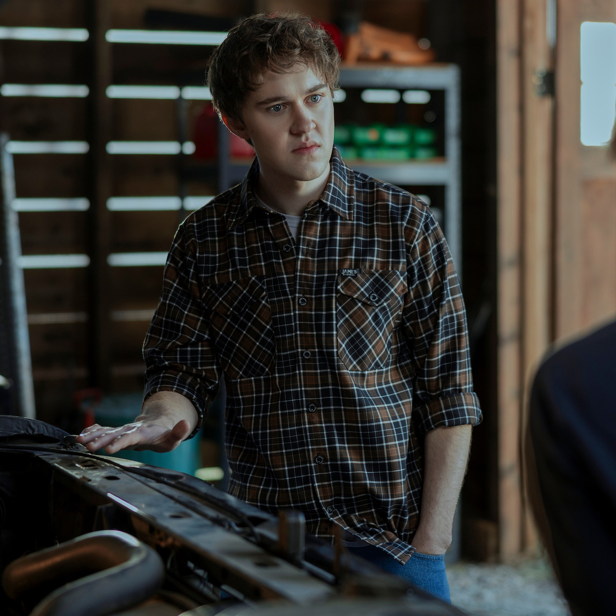 Young man in plaid shirt stands in a garage or workshop, resting one hand on a car part, with shelves and tools visible in the background, daylight coming in from an open door.