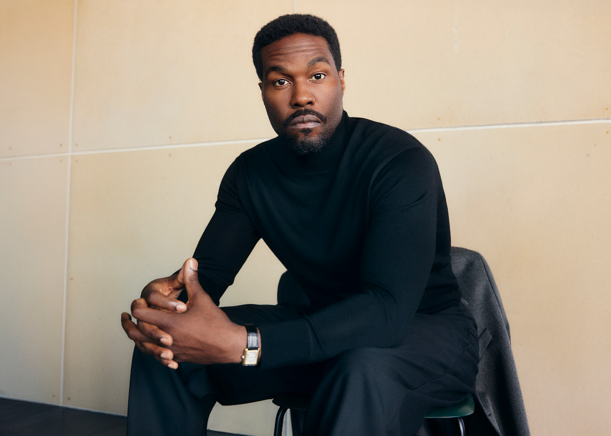 Man in black turtleneck and watch sitting on a chair, hands clasped, with a contemplative expression in a modern, minimalist room with beige walls.