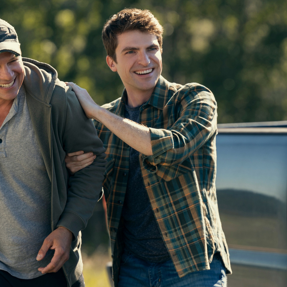Two smiling people outdoors, one with a plaid shirt, standing close together by a vehicle with trees and sunlight in the background, appearing relaxed and happy.