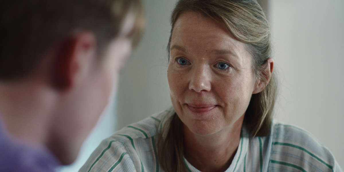 A woman with light brown hair and a striped shirt smiles gently while sitting indoors, facing a person whose face is partially visible in the foreground.