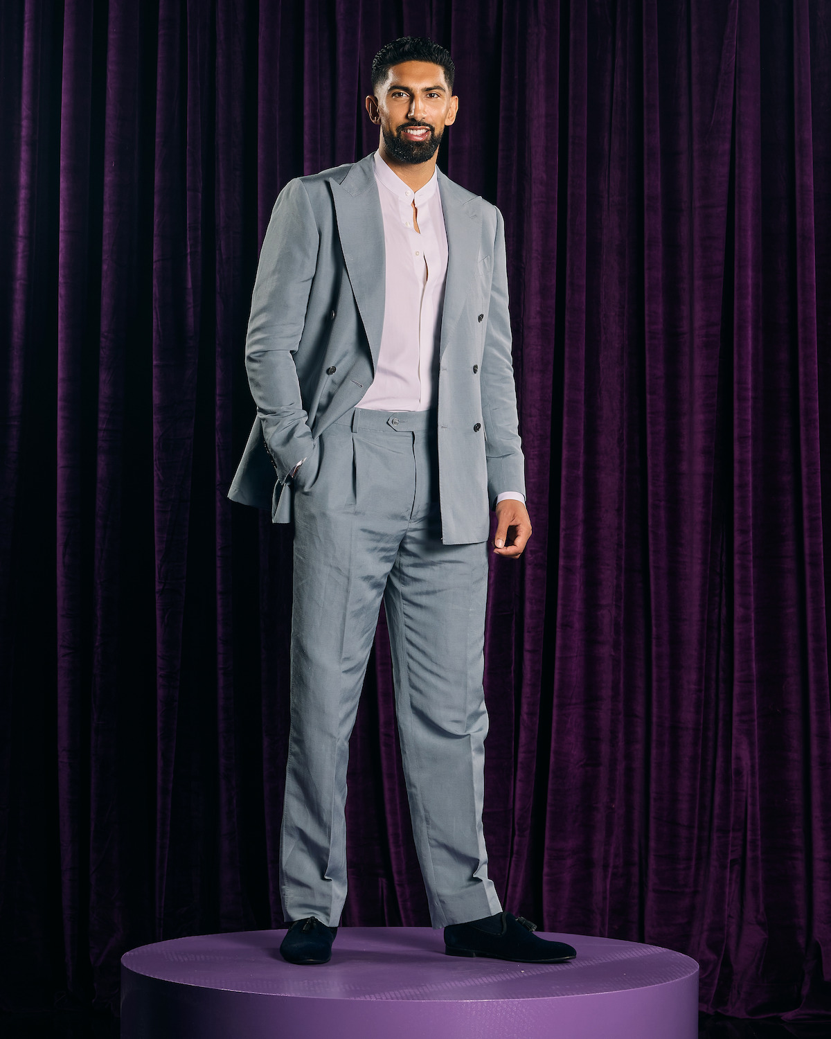 Man in a light gray suit with a white shirt stands confidently on a purple circular platform, in front of a background of dark purple velvet curtains, in a studio setting with professional lighting.