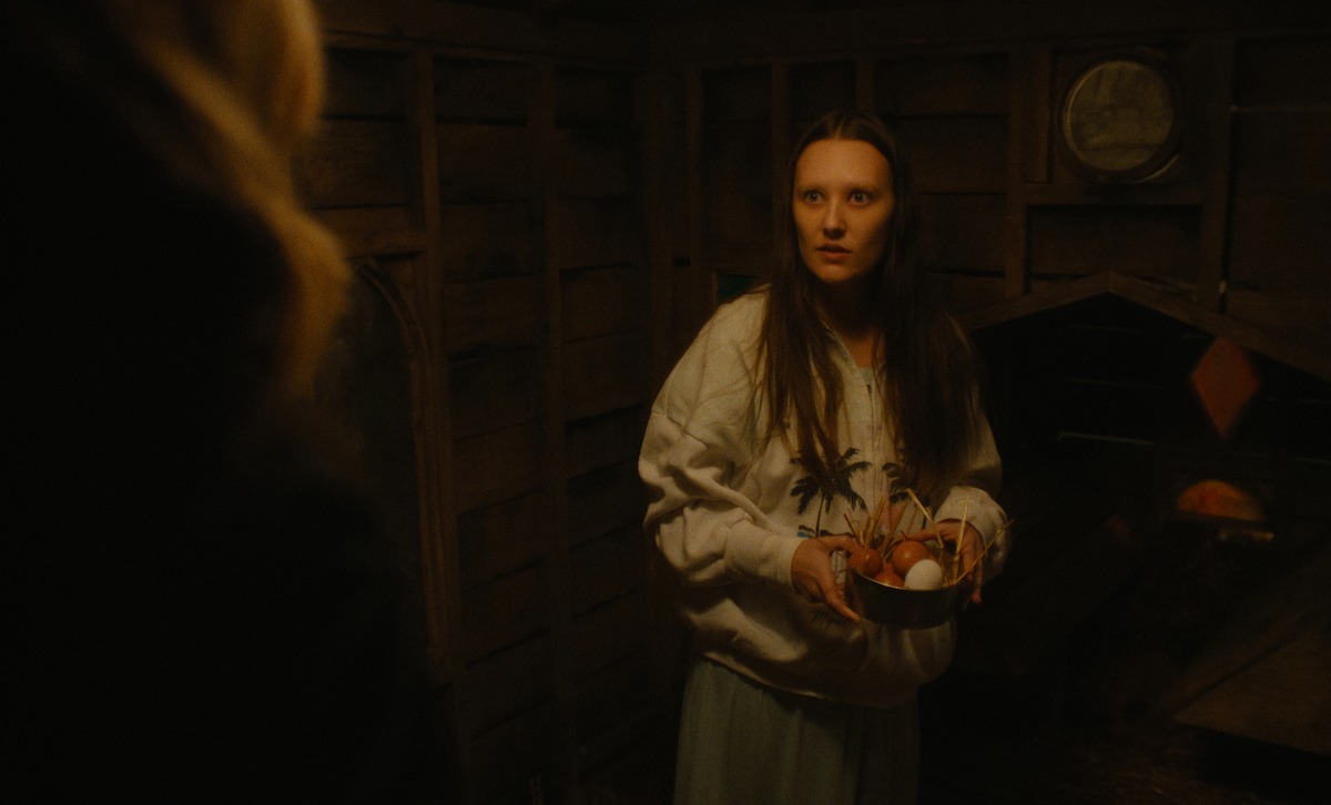 Woman in a dimly lit wooden shed holding a wire basket with fresh eggs, wearing a light sweatshirt and looking at a person in the foreground, rustic interior with wooden plank walls and coop section in background.