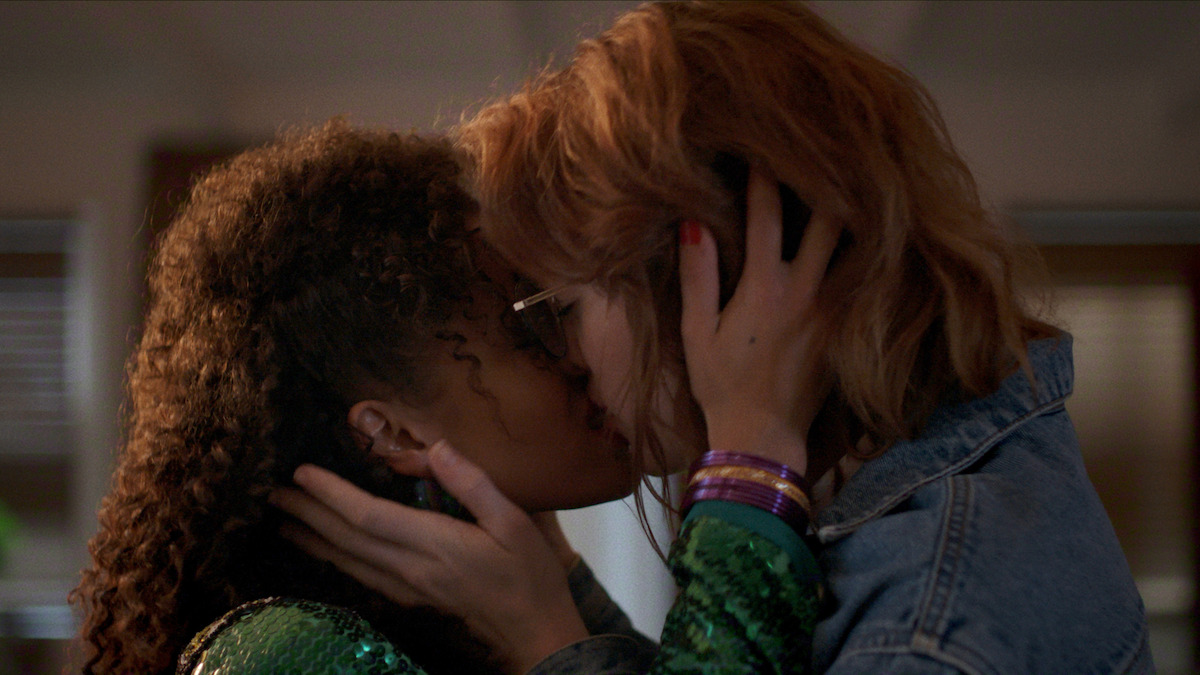 Two women are sharing a romantic kiss indoors, gently holding each other's faces. The background is softly lit, suggesting an intimate, private setting.
