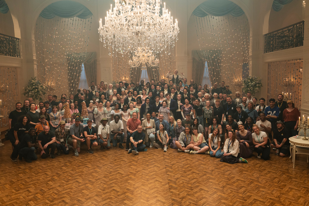 Large group of people gathered for a photo in an elegant ballroom with chandeliers, wooden parquet floor, floral decorations, and draped windows, suggesting an event or celebration in a formal or historic setting.