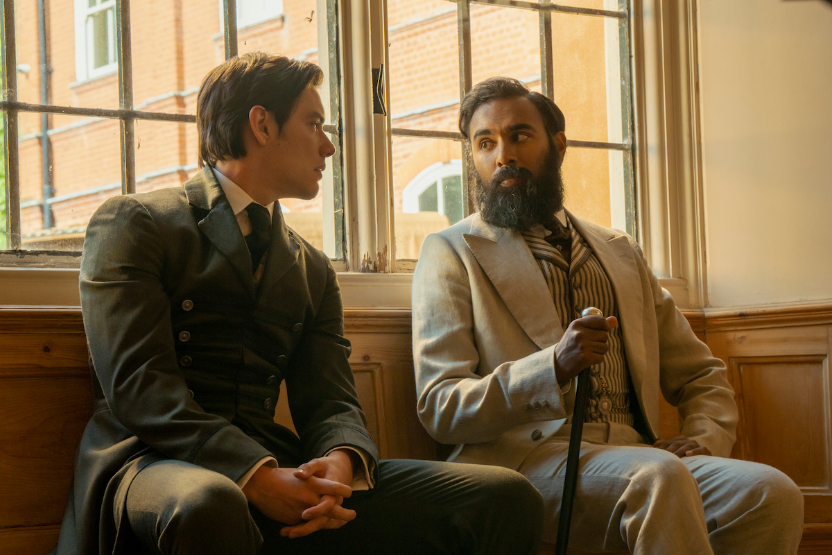 Two men in period clothing sit on a wooden bench by a large window in a sunlit room, engaged in conversation. One wears a dark suit, the other a light suit and holds a cane, with brick buildings visible outside the window.
