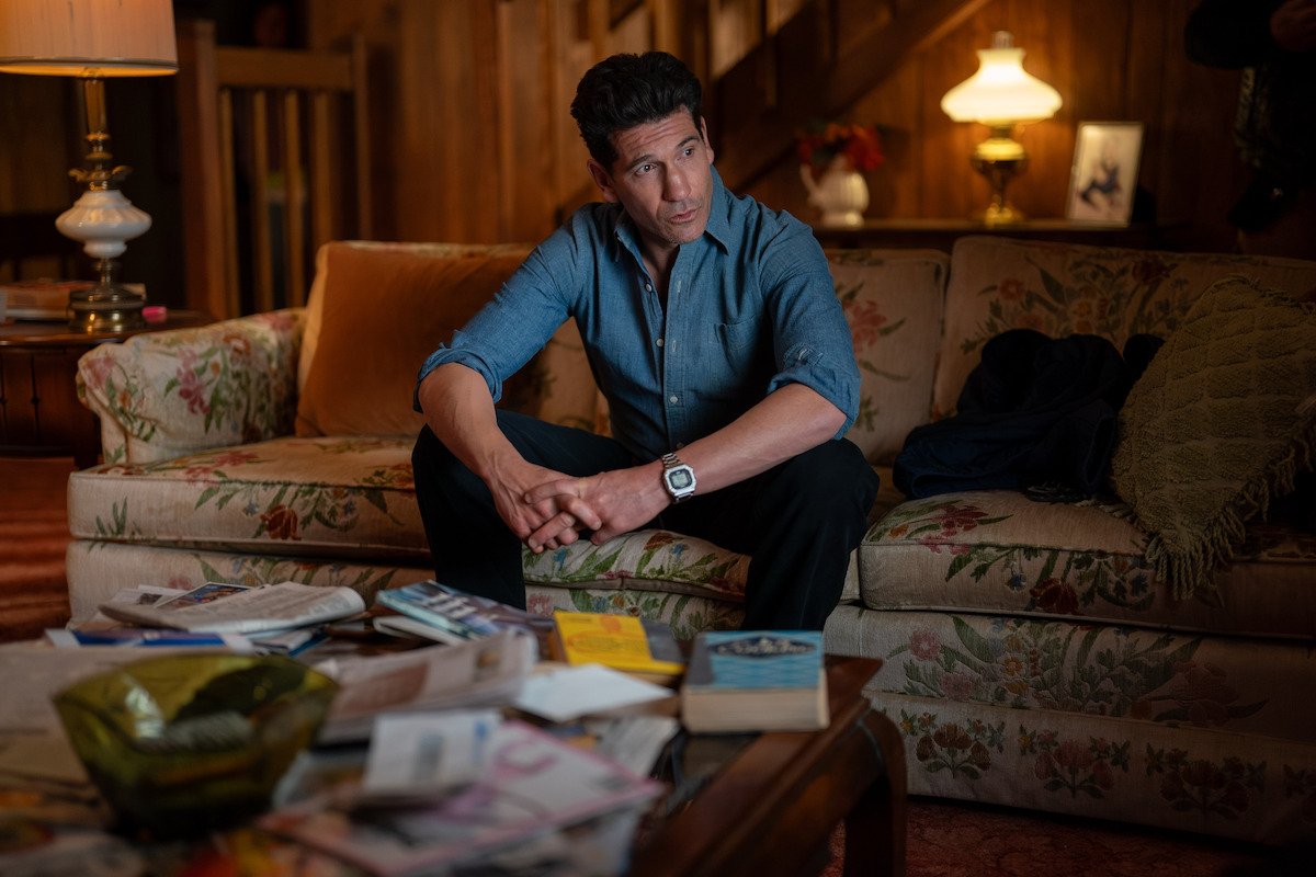 Man sits on a floral sofa in wood-paneled living room. Coffee table cluttered with books and papers.