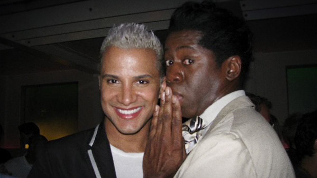 Two men in dressy attire pose indoors; one is smiling at the camera with platinum blond hair, while the other, wearing a bow tie, leans in to whisper with a playful expression. Dimly lit room with people and neutral colors in background.