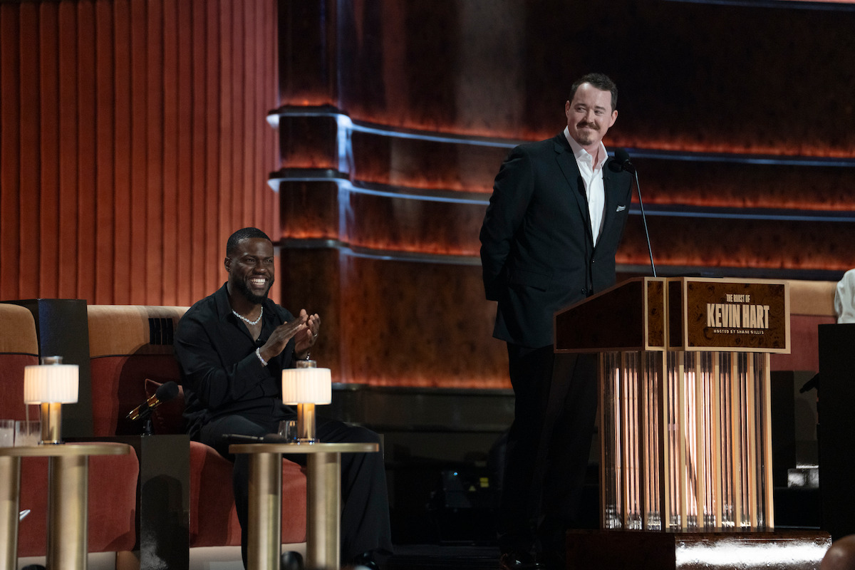 Two men on stage at a comedy roast, one sitting and clapping, the other standing and speaking at a podium labeled "Kevin Hart." The setting has warm lighting with elegant furniture and a formal, theatrical backdrop.