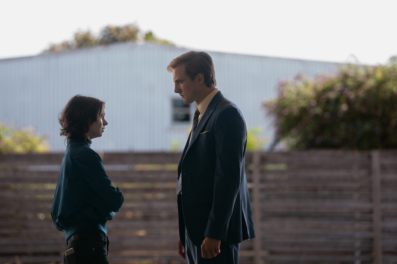 Two people stand facing each other outdoors in front of a wooden fence and a metal building, having a serious conversation in daylight.