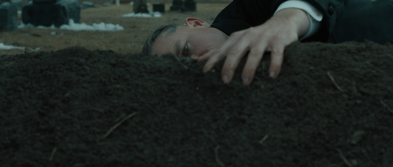 A person in formal clothing lies on the ground in a cemetery, reaching out and touching a mound of dirt with a somber expression.