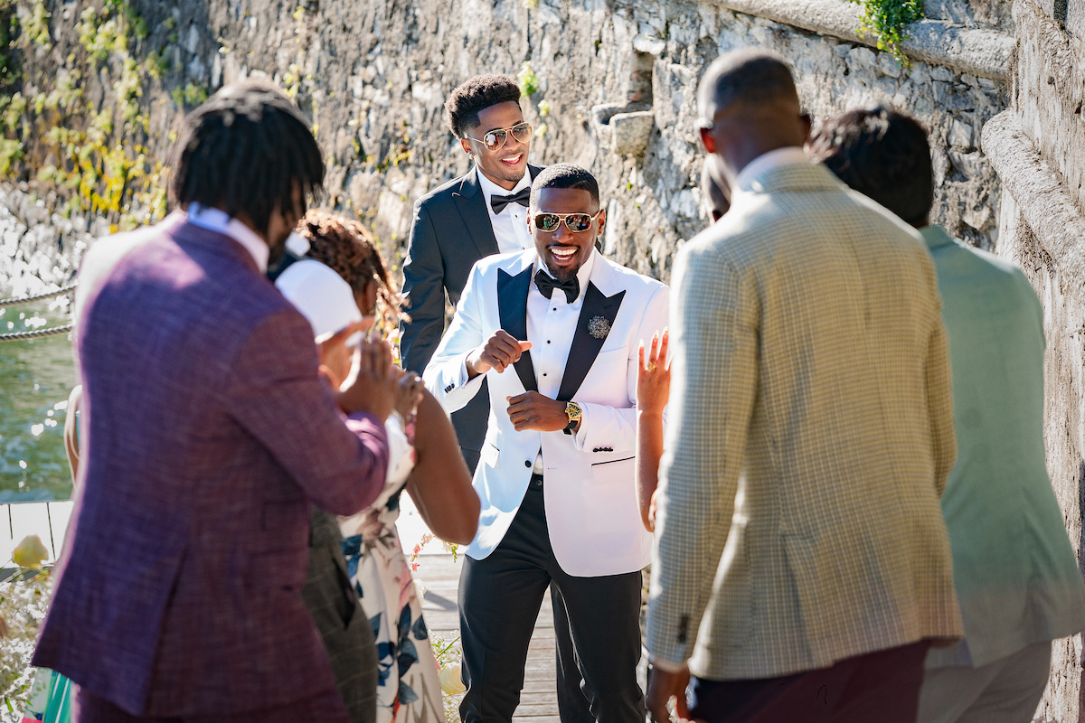 A joyful man in a white tuxedo jacket walks towards friends outdoors near a rocky wall, smiling and celebrating at a formal event, possibly a wedding or special occasion.