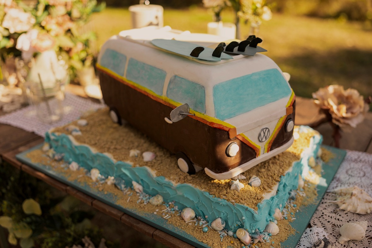 A cake shaped like a vintage VW van with surfboards on top, decorated to look like it’s on sand and water, surrounded by shells, set outdoors on a rustic wooden table with flowers and sunlight in the background.