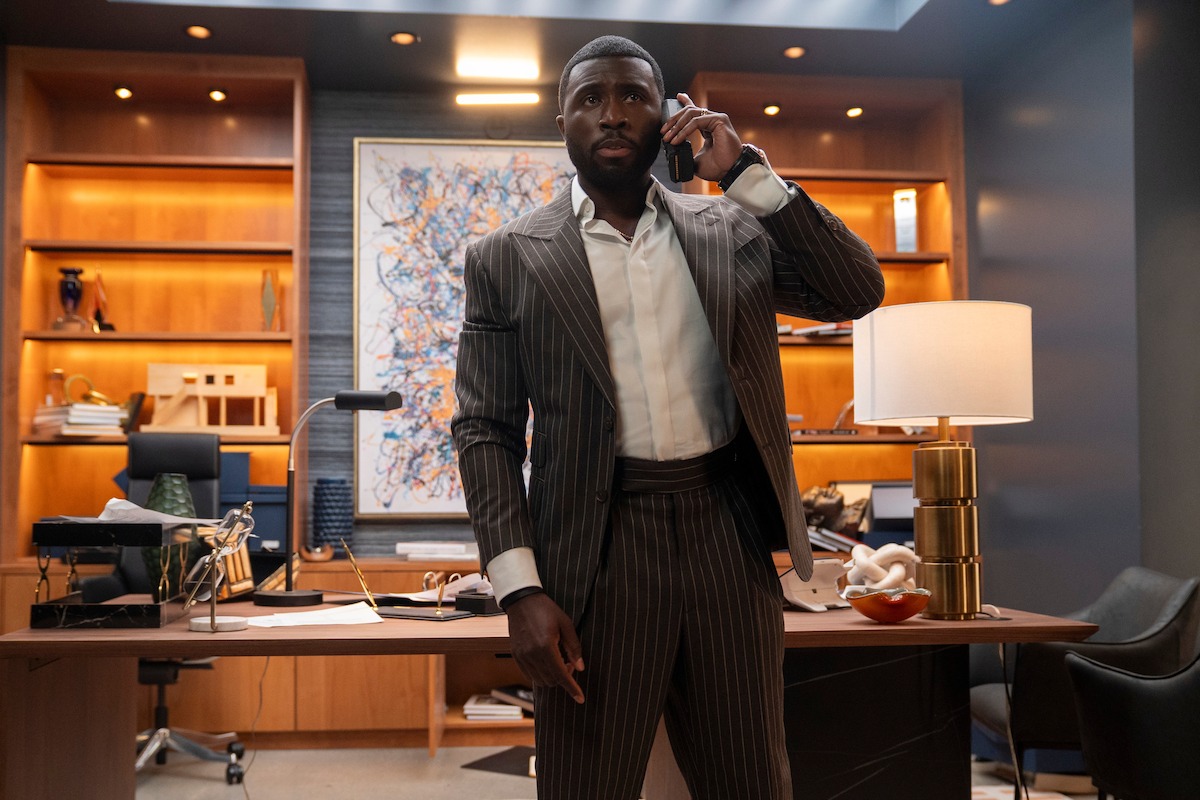 Man in a pinstripe suit stands in a modern, well-lit office, speaking on a phone. Bookshelves, a large abstract painting, desk lamp, and organized workspace are visible in the background, conveying a professional environment.