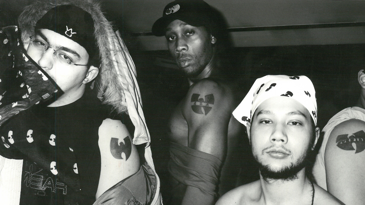 Three men pose in a black and white indoor photo, showing off Wu-Tang Clan tattoos and clothing featuring the group's logo. The environment is casual with urban streetwear and bandanas, emphasizing their affiliation and attitude.