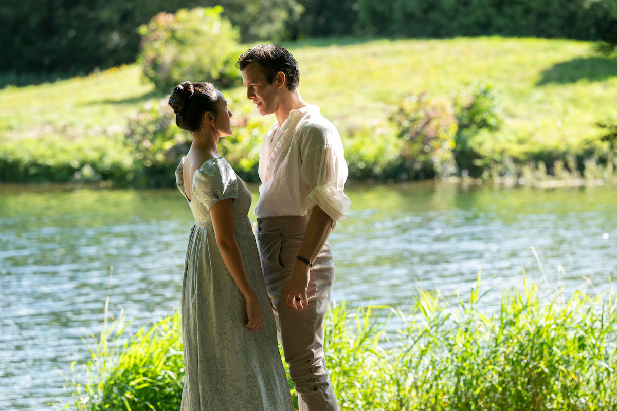 A man and woman in period clothing stand closely facing each other by a riverside, surrounded by green grass and bushes on a sunny day.