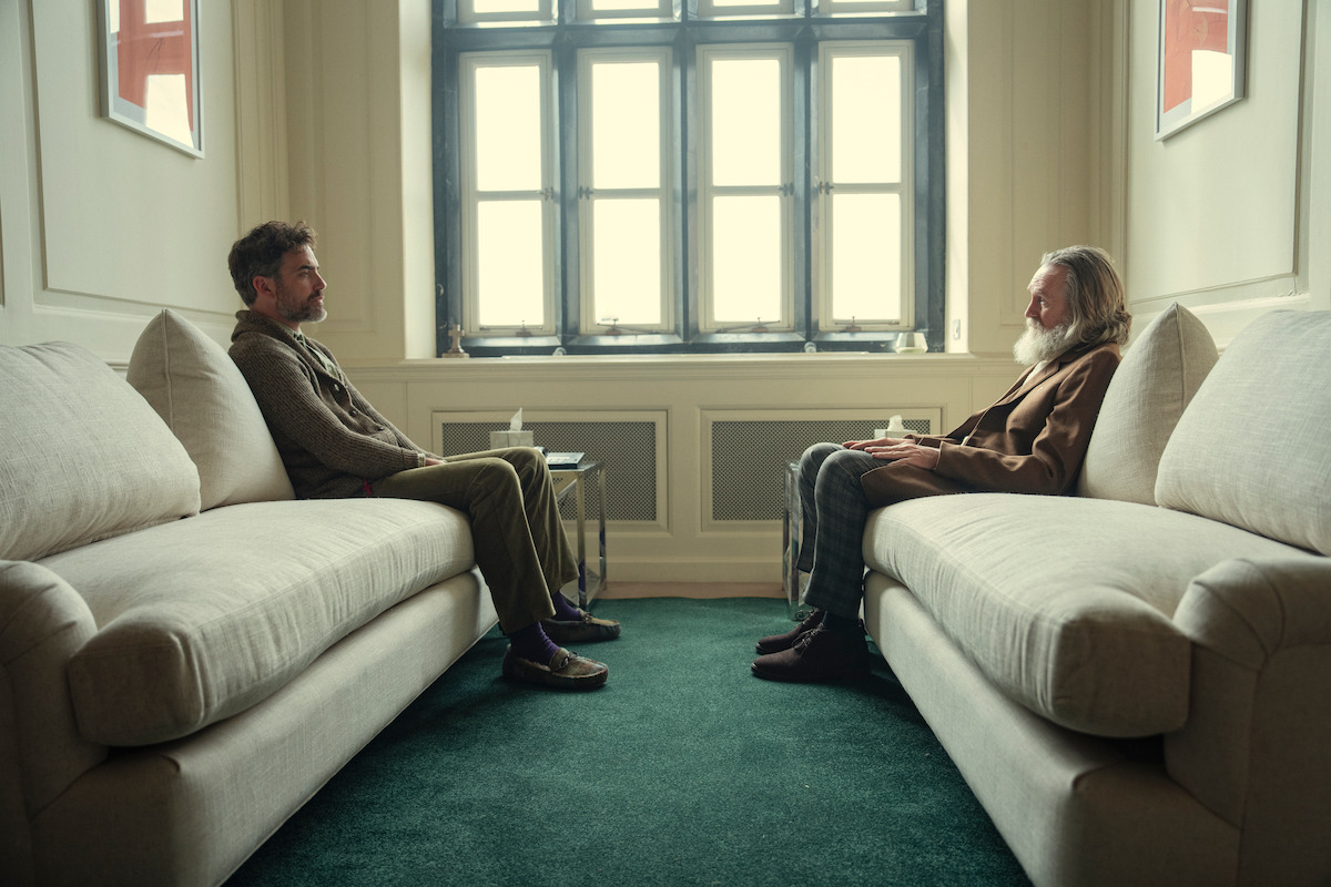 Two men sitting across from each other on separate couches in a softly lit room with a large window, green carpet, and neutral decor, appearing to be in a thoughtful or conversational setting.