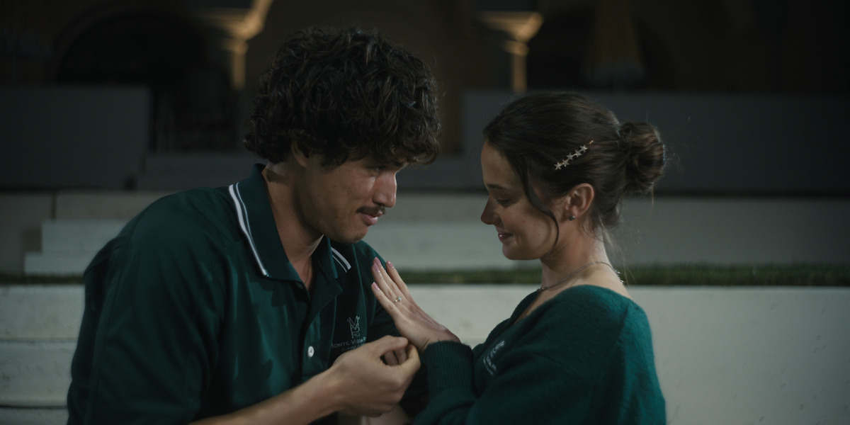 A young man and woman stand close together outdoors at night, both wearing green collared shirts, sharing an intimate moment and looking at each other’s hands, with blurred background arches suggesting a quiet, possibly romantic setting.
