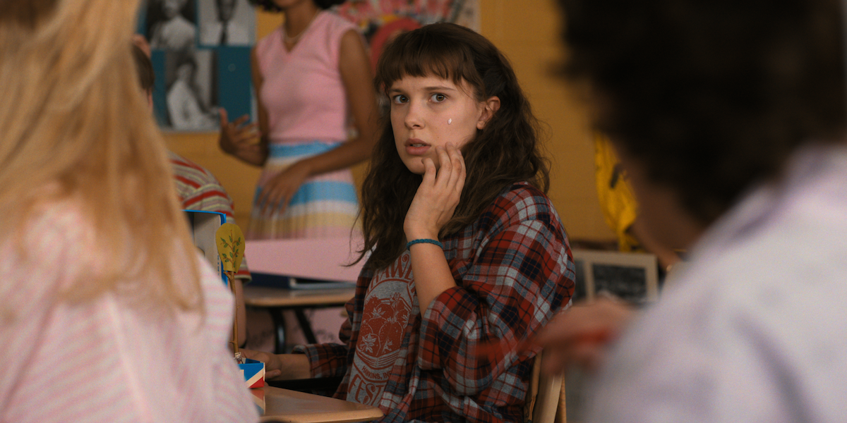 A teenage girl in a plaid shirt sits in a classroom, looking surprised and touching her face.
