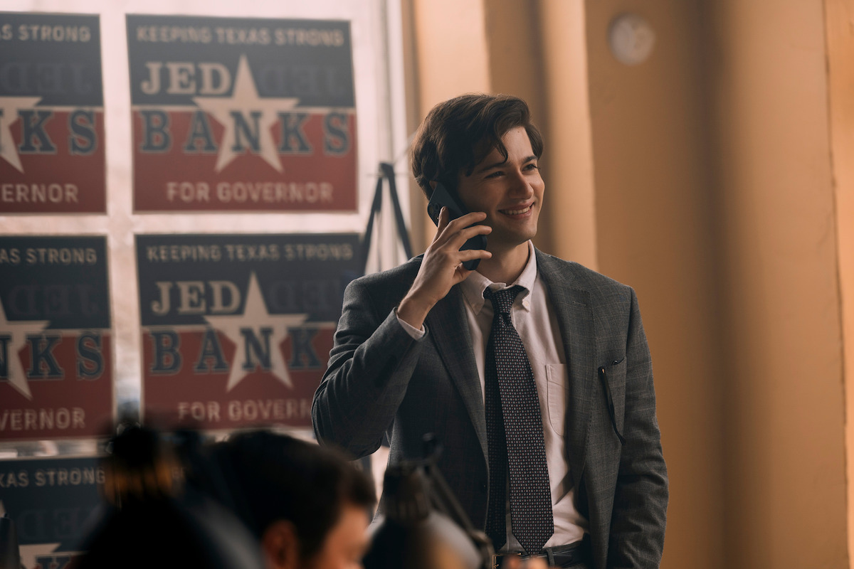Young man in a suit smiling while talking on a phone in an office decorated with political campaign posters that read "Jed Banks for Governor" and "Keeping Texas Strong." Warm indoor lighting with other people working in the background.