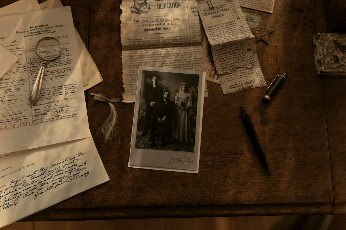 Old sepia photograph, handwritten letters, magnifying glass, pen, and newspaper clippings spread out on a wooden desk, suggesting investigation or family history research in a vintage setting.