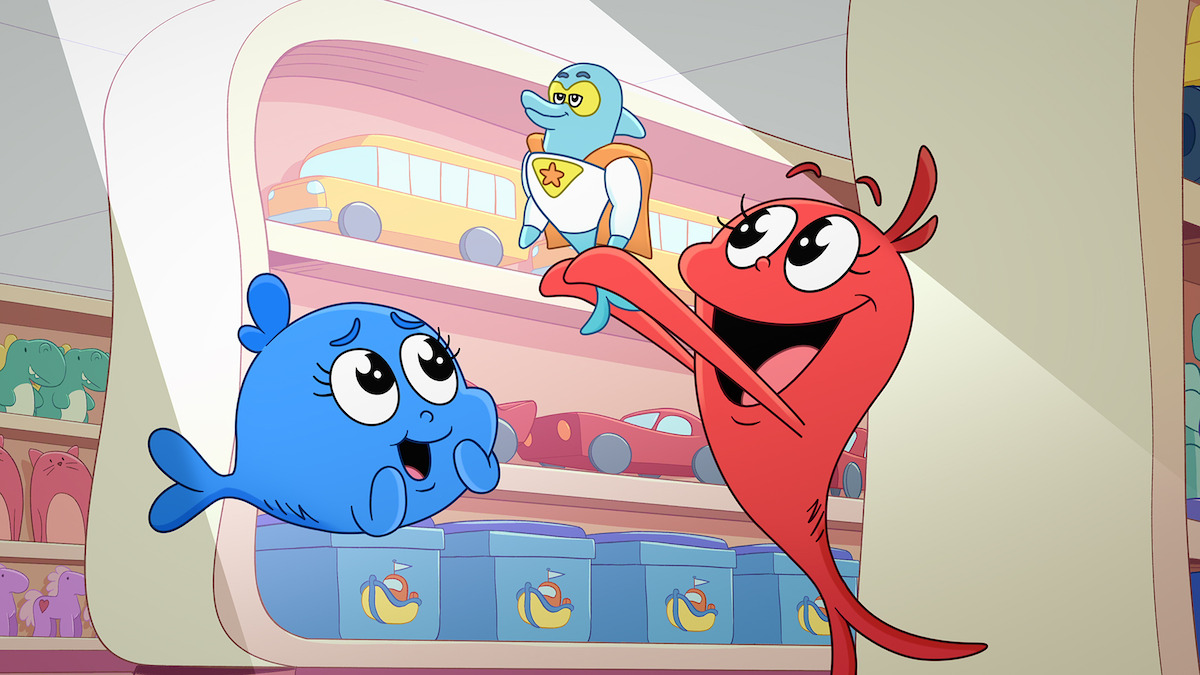 Two cartoon characters, a blue fish and a red heart, are inside a toy store. The red heart excitedly holds up a superhero duck toy while the blue fish watches. Shelves with various toys appear in the background.