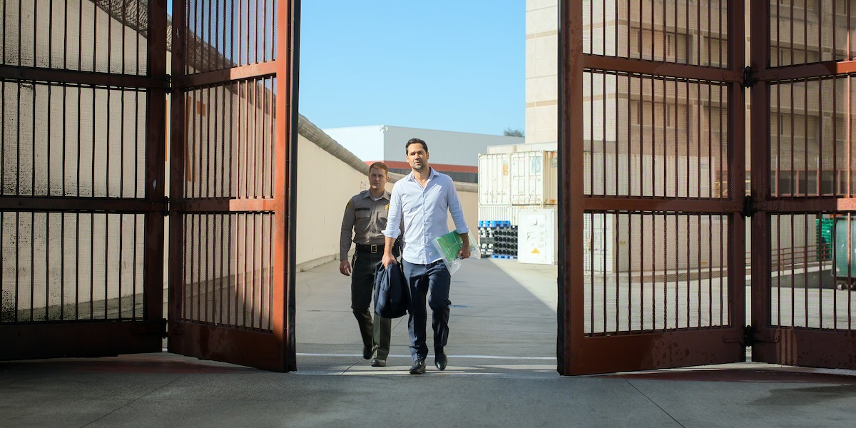 A man carrying a duffel bag and folder walks out of large metal gates in a sunny outdoor facility, accompanied by a uniformed officer, suggesting release from a secure or institutional environment.