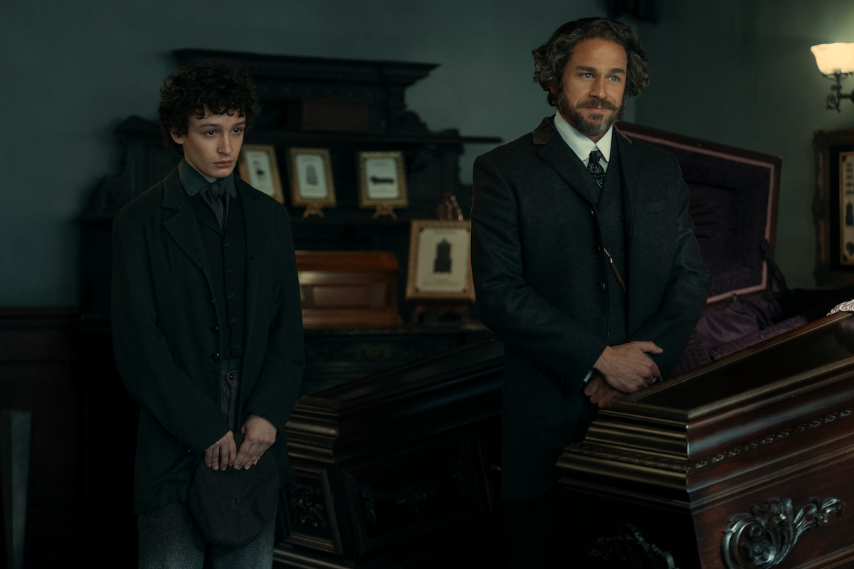 Two men in dark, old-fashioned suits stand beside a polished wooden coffin in a dimly lit, ornate room decorated with framed photos, suggesting a 19th-century funeral or wake setting.