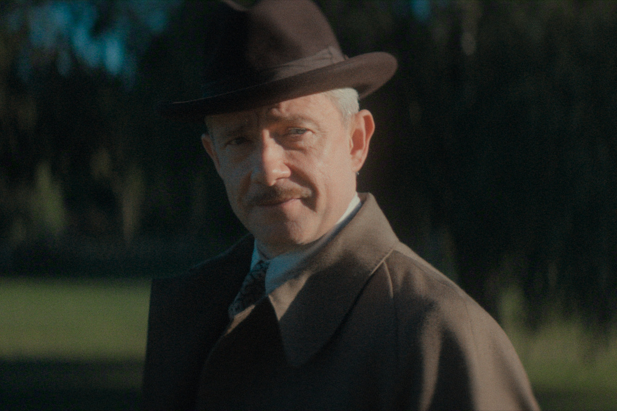 A man with a mustache wearing a dark fedora and brown overcoat stands outdoors in a sunlit, grassy park with trees blurred in the background.