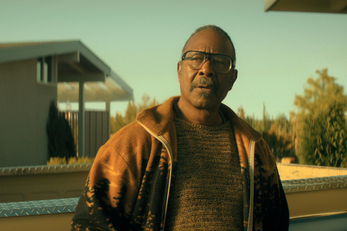 Clarke Peters as Art, with glasses wearing a knit sweater and orange jacket stands outdoors in a residential area, with modern houses and greenery visible in the background on a sunny day.