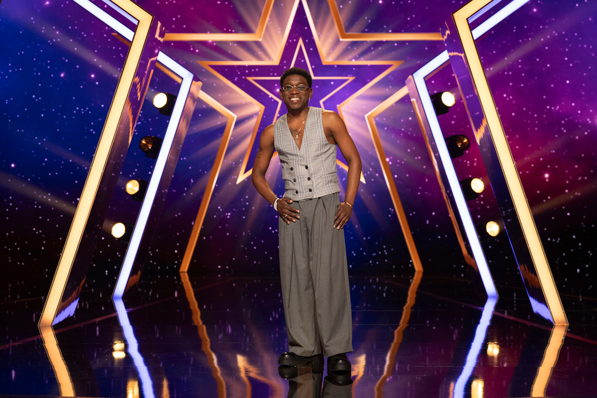 Steven “Spud” Howard standing center stage in front of a large illuminated star, wearing a vest and wide-legged pants, with a vibrant purple and gold background and stage lights, creating a show or competition atmosphere.